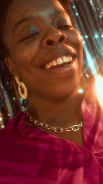 Doğum günü pastasında dilek tutan ve mum üfleyen mutlu Afrikalı genç bir kadının dikey yavaş çekimi, partideki parlak folyo folyo perde duvarına karşı duruyor.