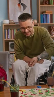 Evdeki rahat oturma odasında arkadaşlarıyla masa oyunu oynarken tekerlekli sandalyede eğlenen neşeli beyaz adamın dikey görüntüsü.