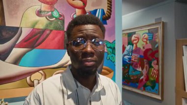 Modern sanat galerisindeki duvarda asılı duran ve kameraya bakan beyaz gömlekli ve gözlüklü genç Afrikalı Amerikalı adamın portresini kapat.