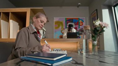 Modern sanat galerisinde masa başında oturan, not defterinde yazan ve arka planda dizüstü bilgisayarında çalışan siyahi bir erkek meslektaşla günlük planlamacıya bakan yetişkin, beyaz bir kadın küratör.