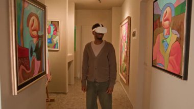 Gelişmiş gerçeklikteki soyut sanat sergisini keşfederken VR kulaklıklı genç Afrikalı Amerikalı erkek ziyaretçinin orta boy uzun bir görüntüsü.