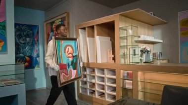 Genç Afrikalı Amerikalı erkek galerinin, çerçeveli, soyut bir resim getirip modern sanat galerisindeki sergiye hazırlanırken onu resepsiyon masasına koyarkenki görüntüsü.
