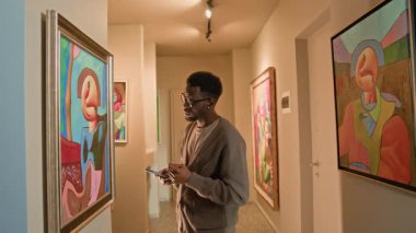 Orta boy Afro-Amerikan bir adamın sergi sırasında soyut resim gözlemlemesi kulaklıklarda müzik rehberinin galeride sergilenen sanat eserlerini öğrenmesi.
