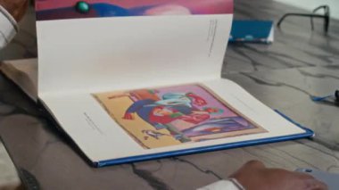 Siyahi adamın, modern sanat galerisinde önünde oturan olgun, beyaz bir kadınla yaptığı sohbete katılan baskılı resimlerle sayfalarca resim çevirdiği el resmi.