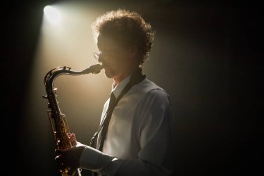 Odaklanmış gözlüklü bir erkek saksafoncunun karanlık stüdyoyu büyüleyici müziklerle doldurup dikkatle rüzgar enstrümanı çalması.