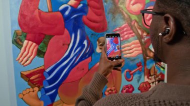 Genç Afrikalı Amerikalı erkek galeri ziyaretçisinin akıllı telefon kamerasıyla soyut resim çekerken telli kulaklıklarda ses rehberini dinlerken çekilmiş yavaş çekimleri.