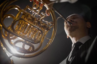 Düşük açılı, Fransız kornosu çalan erkek müzisyenin gözleri kapalı sahnede parlak spot ışıklarıyla aydınlatılmış büyüleyici bir müzik çalarken.
