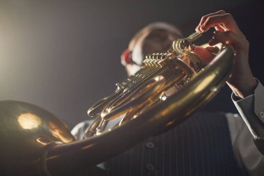 Düşük açılı çekimde, erkek müzisyen altın Fransız kornosu valf kollarını iterken sahnedeki parlak spot ışıkları altında derin ve yumuşak bir müzik icra ediyor.
