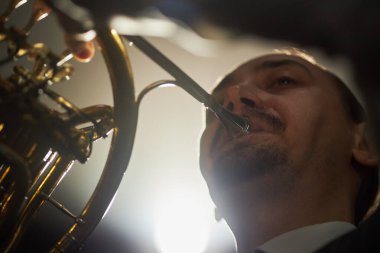 Fransız kornosu çalan profesyonel erkek orkestra müzisyenine yakından bakın. Parlak spot ışıkları altında sahnede müzik icra ediyor.