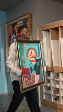 Genç Afrikalı Amerikalı erkek galericinin dikey görüntüsü çerçeveli bir resim getiriyor ve modern sanat galerisinde sergi hazırlarken onu resepsiyon masasına koyuyor.