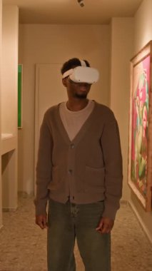 VR kulaklıklı genç Afrikalı Amerikalı erkek galeri ziyaretçisinin dikey orta boy fotoğrafı. Arttırılmış gerçeklikte sanat sergisi keşfederken etrafa bakınıyor.