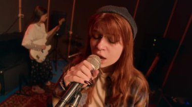Arkadaşları müzik stüdyosunda elektro gitar ve davul çalarken, kızıl saçlı ve bereli bir kadının mikrofona şarkı söylediği yüksek açılı bir portre çekimi.