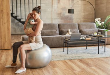 Özürlü bir annenin tam boy portresi. Bebeği kucağında tutarken rahat evdeki fitness topunda zıplayıp duruyor. Boşluğu kopyala.