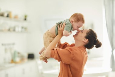 Mutlu genç annenin belden yukarı portresi. Sıcak bir evde bebekle oynuyor. Güneş ışığıyla aydınlatılıyor, boşluğu kopyalıyor.