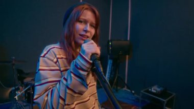 Çizgili süveter giyen, rock şarkısını mikrofona söyleyip müzik stüdyosundaki kameraya bakan kızıl saçlı punk kızın portresini çekiyorum.