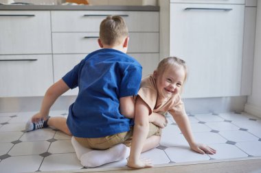 Down sendromlu mutlu küçük kızın uzun bir portresi mutfakta erkek kardeşiyle oynuyor ve kameraya bakıyor, boşluğu kopyalıyor.