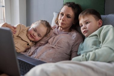 Değer veren annenin portresi kanepeye uzanmış, dizüstü bilgisayarla video izlerken Down sendromlu oğlunu ve kızını kucaklıyor.