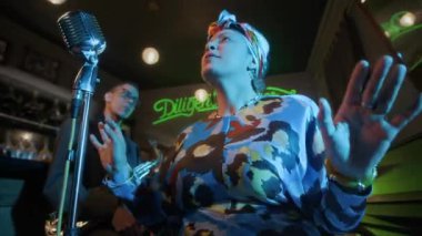 Enerjik çift ırklı kadın caz vokalisti, klasik mikrofona doğru şarkı söylerken, bardaki müzisyenlerle yeşil neon ışıklı canlı performans sergiliyor.