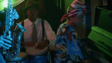 Caz üçlüsü neon ışıkları altında gösteri yapıyor. Ön planda saksafoncu, merkezde conga çalan perküsyoncu ve renkli giyinen kadın piyanist neon barda canlı, ritmik bir sahne yaratıyor.