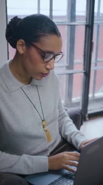 Modern ofiste dizüstü bilgisayarda çalışan gözlüklü siyahi iş kadınının dikey yavaş çekimi.