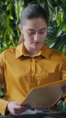 Modern yeşil çalışma sahasında oturmuş kağıt okuyan, sonra da kameraya gülümseyen beyaz iş kadınının dikey orta portresi.