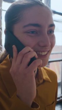 Pencerenin önünde telefonla konuşan genç, neşeli, beyaz bir kadının dikey yakın çekimi.