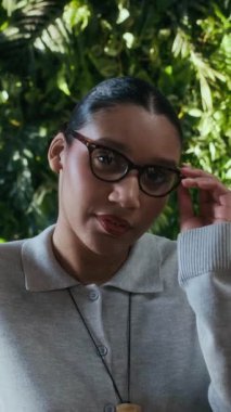 Afro-Amerikan bir iş kadınının portresindeki dikey zum. Formalite giysisi içinde, gözlük takıyor ve yemyeşil duvarın önünde kameraya gülümsüyor.