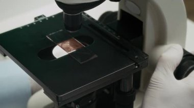Beyaz eldivenli, yoğunlaşmış gri saçlı yaşlı bilim adamının araştırma laboratuarında mikroskopta cam kaydırma testine baktığı yavaş çekim.