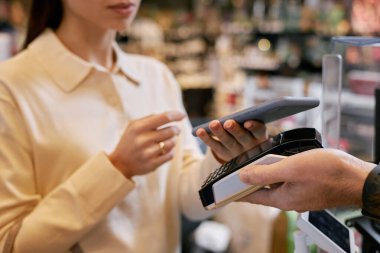 Kasiyer ödeme terminalini tutarken bir kadın dijital ödeme işlemleri için telefon kullanıyor. Arka plandaki bulanık ürünler perakende ortamı hissi yaratıyor