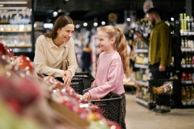 Anne ve kızı bir süpermarkette taze ürünleri incelerken gülümsüyor ve sohbet ediyorlar. Etrafı taze meyvelerle çevrili.