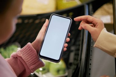 Arka planda meyve ve sebzelerle süpermarkette alışveriş yaparken başka birine akıllı telefon ekranı gösteren kişinin yakın plan görüntüsü