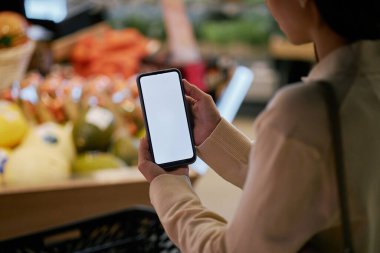 Markette alışveriş yaparken akıllı telefon tutan, meyve ve sebze bölümündeki ürünleri inceleyen, cihaz üzerindeki bilgileri kontrol eden bir kadın.