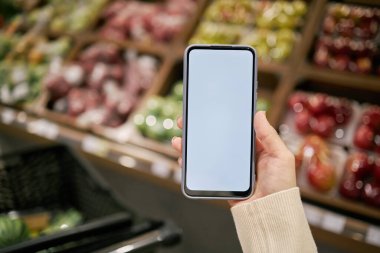 Markette arka planda raflarda taze ürünler sergilenen akıllı telefonu tutan insan eli. Akıllı telefonun boş ekranı dizayn veya metin eklemek için kullanılabilir