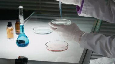 Orta yaşlı, koruyucu gözlüklü ve eldivenli beyaz kadın bilim adamının, petri kabından küçük miktarda sıvı transferini ve incelenen maddeyi yakından çek.