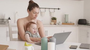 Beyaz tenli bir iş kadınının mutfak masasında kucağında oğlunun laptopuyla oturduğu orta boy bir fotoğraf.