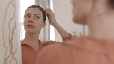 Beyaz saçlı, siyah saçlı, güzel, orta yaşlı bir kadının aynadaki yansımasına baktığının arka görüntüsü.