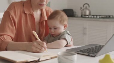 Orta büyüklükte, beyaz kadın, dizüstü bilgisayarın önünde dizlerinin üstüne çöküp not defterine notlar yazarken çekilmiş.