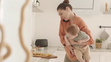 Kafkasyalı güzel bir kadının elinde bebekle mutfak atölyesinde cep telefonuyla sohbet ederken kahvaltı hazırladığı orta boy bir fotoğraf.