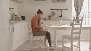 Orta yaşlı beyaz kadın bacak protezi takmış, kucağında bebekle yemek masasında oturuyor ve onu besliyor.