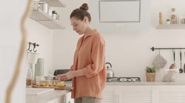 Orta yaşlı beyaz bir kadının mutfak atölyesinde kahvaltı hazırlarken ön planda güvenli bebek koltuğunda yatıp onu sakinleştirmeye çalışırken çekilmiş orta boy bir fotoğrafı.