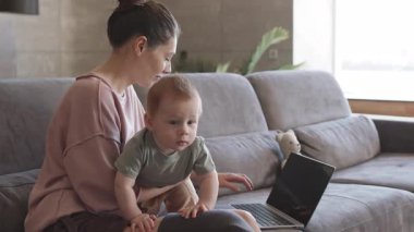 Orta yaşlı, beyaz, bacak protezi takmış bir kadının oturma odasında kucağında kucağında bebekle dizüstü bilgisayarla oturduğu bir fotoğraf.
