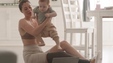 Geniş açılı güzel orta yaşlı beyaz kadın bacak protezi ile mutfak zemininde oturuyor ve elinde çocuğu ve video çağrısı var.