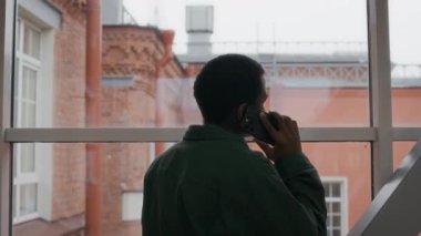 Orta boy siyahi bir çocuğun koridorda büyük pencerenin önünde telefonla konuşurken, kamerayı çekerken, boşluğu kopyalarken görüntüsü.