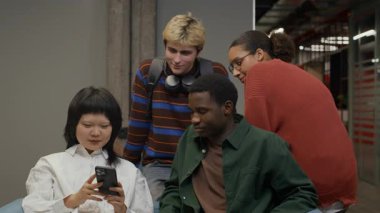 Orta ırklı bir grup genç öğrenci telefonda komik videolar izliyorlar. Üniversite koridorundaki kanepede dinleniyorlar. Çalışma saatleri arasında eğleniyorlar.