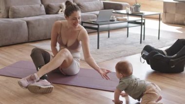 Antrenmandan sonra oturma odasında lastik paspasın üzerinde oturan ve yanında emekleyen bebekle oynayan sportif bir anne resmi.