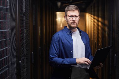 Sunucu odasında sunucuları yönetirken elinde laptop tutan gözlüklü bir adamın portresi. Mavi gömlek giyip sunucu performansını analiz etmek