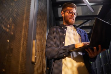 Gözlüklü bir adam sunucu odasında sunucu ışığıyla aydınlatılmış bir dizüstü bilgisayarla çalışıyor. Sıradan bir tişört giyerken sunucu raflarının yanında duruyor.