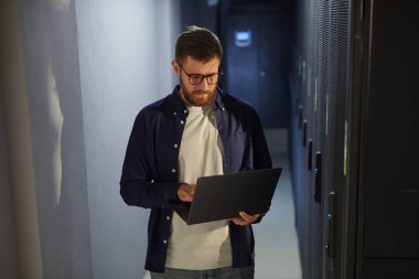 Bir veri merkezinin karanlık koridorunda sakallı ve gözlüklü bir adam sunucu raflarını ve ekipmanlarını incelerken dizüstü bilgisayarda çalışıyor.