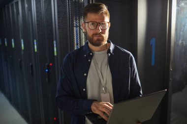 Sakallı, gözlüklü ve dizüstü bilgisayarlı bir adamın portresi sunucu rafları ve loş ışıklandırmalı bir sunucu odasında profesyonel ve odaklanmış bir atmosfer yaratıyor.