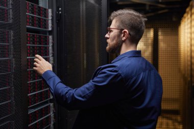 Modern veri merkezindeki sunucu donanımını ayarlayan teknisyen görülebilir raflarla birlikte. Mavi tişört giyen kişi, göreve odaklanırken yan görünüm takınıyor